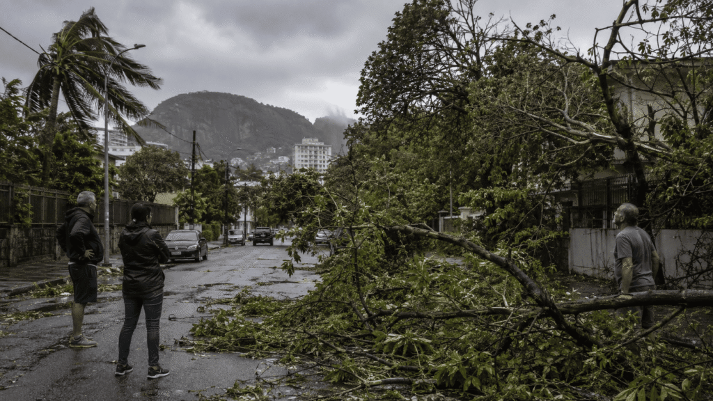 Árvores e galhos caídos bloqueiam uma rua molhada no Rio de Janeiro após fortes rajadas de vento, enquanto moradores observam os danos.